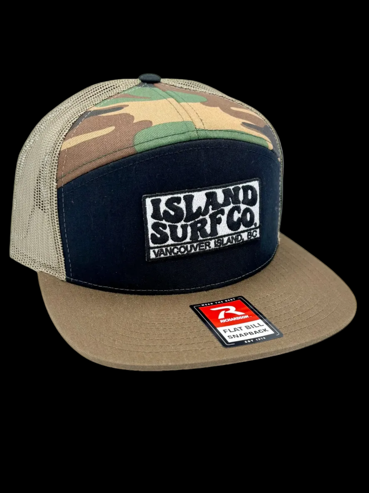 A trucker-style snapback cap with a camouflage pattern on the front panels, a dark navy blue front panel, and a tan flat brim. The front features a white embroidered patch reading 'ISLAND SURFCO.